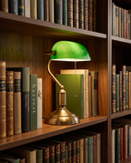 Green Shade Banker's Desk Lamp, Antique Brass LED — on a home library shelf among hardcover books in a warm evening glow